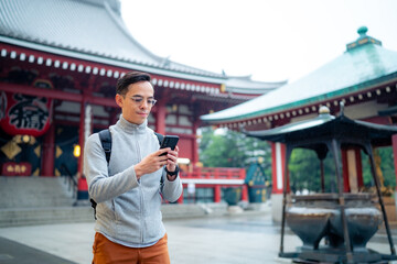 Hispanic tourist guy standing on blurred background of Japanese temple Sensoji  and sending text message via mobile phone during journey in Asakusa Tokyo, Japan