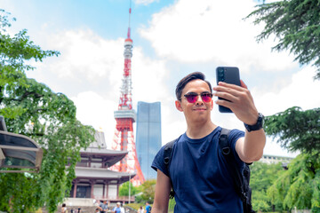 Smiling young Hispanic male traveler in casual wear and sunglasses smiling and taking selfie with smartphone while standing against ancient Zojo ji Buddhist temple near Tokyo tower, Japan