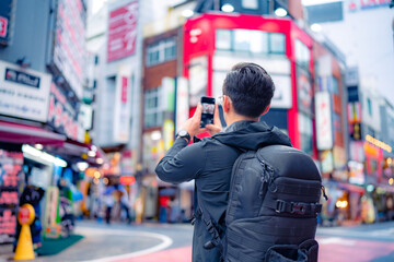 Back view of unrecognizable Hispanic male tourist with backpack taking picture on smartphone in bright Shinjuku neighborhood street of Tokyo city, Japan - obrazy, fototapety, plakaty