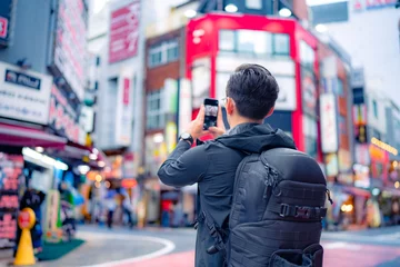 Poster Back view of unrecognizable Hispanic male tourist with backpack taking picture on smartphone in bright Shinjuku neighborhood street of Tokyo city, Japan © Itza