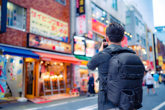 Back view of unrecognizable Hispanic male tourist with backpack taking picture on smartphone in bright Shinjuku neighborhood street of Tokyo city, Japan