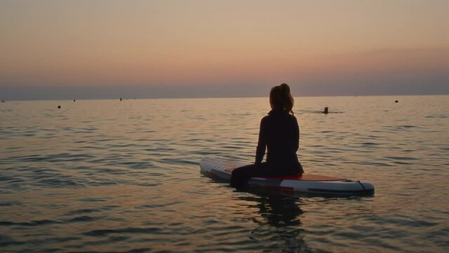 An elderly woman rides paddleboarding in the ocean. Senior female is waiting for a wave on a surfboard. An old woman is sitting on a board in the sea at sunset, dressed in a swimming suit for surfing