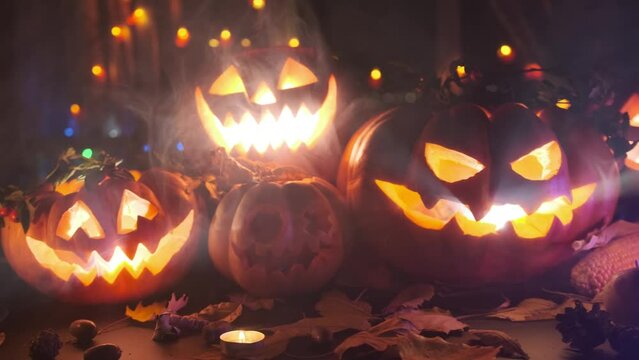 Many creepy pumpkins with scary face carved out, glowing from inside, pumpkins standing along Halloween decorations, flickering candles, cold fog, colorful lights and dry autumn foliage. Close up.