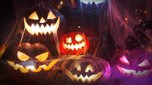 Large pile of traditional jack o'lantern pumpkins in the dark and cobwebs lie on the threshold of the house flashing colorful lights spooky party during Halloween celebrations.