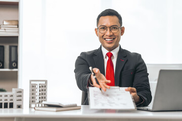 Agent presents contracts for property purchase or lease. Businessman asian indian people showcases miniature model home, signifying secure property insurance. Desk scene in a home sales office.