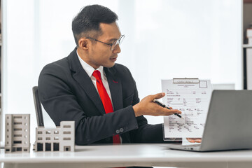 Agent presents contracts for property purchase or lease. Businessman asian indian people showcases miniature model home, signifying secure property insurance. Desk scene in a home sales office.