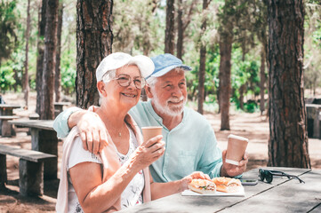 Happy relaxed senior couple enjoying a sandwich while sitting at a wooden table in the woods. Happy elderly man and woman appreciating nature and freedom during retirement