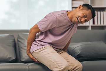Middle-aged Asian Indian man with butt pain, hemorrhoids, colon pain, sitting on a sofa. - obrazy, fototapety, plakaty