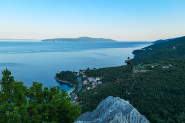 A small metal cross on a rock with a panoramic view of the shore along Medveja, Croatia seen from above. The town is surrounded by thick forest. Endless horizon. An island in the back. Summer holidays