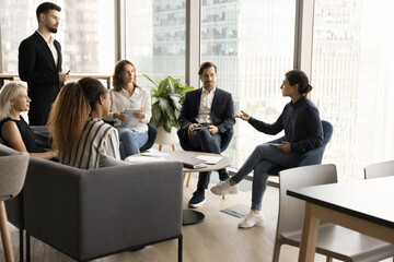 Young Indian business professional speaking on team brainstorming meeting in office lobby, offering ideas for plan, strategy to male boss, talking to diverse group of colleagues