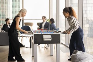 Excited happy diverse colleagues women playing table soccer, having fun at tabletop football game, relaxing in modern office lobby while serious business team talking in background