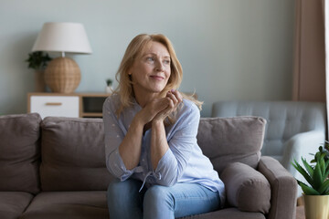 Happy older blonde 50s homeowner woman relaxing on home sofa, thinking, dreaming, looking at window...