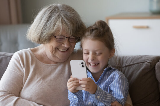 Joyful gen Z granddaughter girl and elder gray haired grandma using smartphone at home together, making video call to family, taking selfie, chatting, enjoying leisure, Internet communication