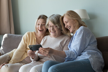 Excited cheerful women of three female generations sharing mobile phone, holding smartphone, resting on home sofa, enjoying leisure time, family meeting, online Internet communication