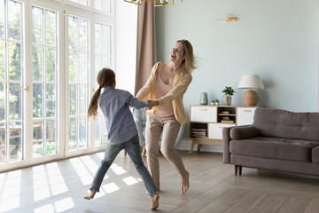 Excited energetic young mom and kid girl holding hands, spinning around, dancing to in living room. Cheerful mother teaching kid to dance at home, smiling, laughing, enjoying family activity
