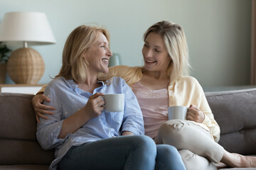 Obraz na płótnie Canvas Happy engaged mature mom and young adult daughter drinking coffee, talking, discussing family life, relatives, laughing, joking, enjoying friendship, leisure time, hugging with love, support