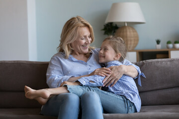 Happy granddaughter girl talking to loving grandma, enjoying trusting friendship, family relations. Caring grandmother embracing grandkid, smiling, laughing, sitting on couch at home