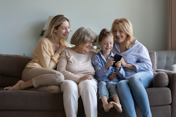 Happy kid, mom, grandma, great grandmother making video call from home couch, smiling, laughing. Child girl holding mobile phone, using online app, playing virtual game with support of family