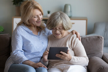 Happy adult mature daughter woman teaching elderly mom to use online app on tablet for communication, hugging elderly lady holding digital gadget, typing, browsing Internet, sitting on home couch - obrazy, fototapety, plakaty