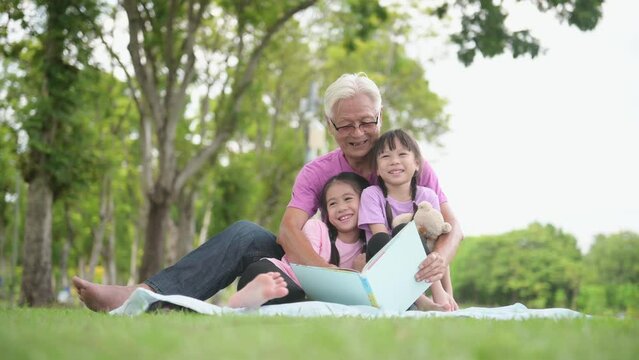 Happy Asian family children reading a book with her grandfather in the park