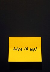A yellow note pad with red written message text Live It Up ,stick on the dark wall - concept of warn oneself to live a happier life while they can
