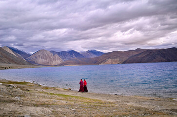 Fototapeta na wymiar Two Buddhist monks taking a stroll on the banks of a lake with blue pristine water under a cloudy overcast morning with a backdrop of mighty Himalayan mountain range