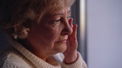 An upset senior woman wiping tears, crying alone near the window, dealing with loneliness in her age