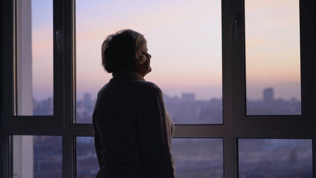 A lonely senior woman sadly looking out the window, missing her family at the retirement home