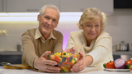Smiling mature spouses showing a bowl with freshly made vegetable salad, healthy nutrition in old age