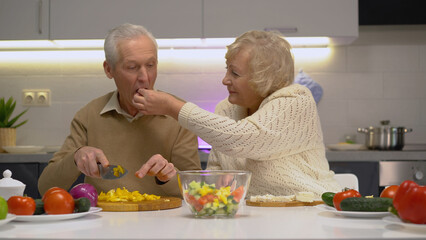 A senior woman treating her husband to tasty cheese while couple is cooking a delicious and healthy meal in the kitchen