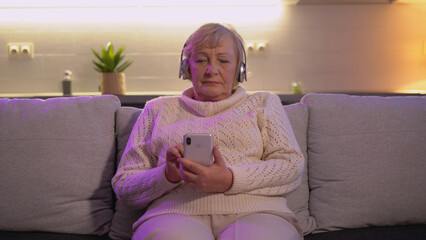 A positive senior woman in wireless headphones is enjoying music and using a phone app