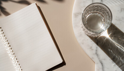 Elegant morning flat lay in a soothing beige palette. A glass of water and an empty notebook rest on a marble table, beautifully illuminated by gentle, dappled sunlight. This minimalist arrangement se - obrazy, fototapety, plakaty