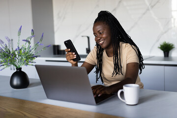 Portrait of young black woman using her smartphone and modern laptop reading text message working at home office - obrazy, fototapety, plakaty