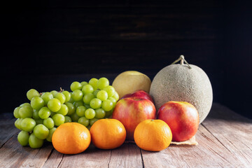 Orange fruits, grapes, apples, cantaloupe placed on a wooden table, black and dark background.