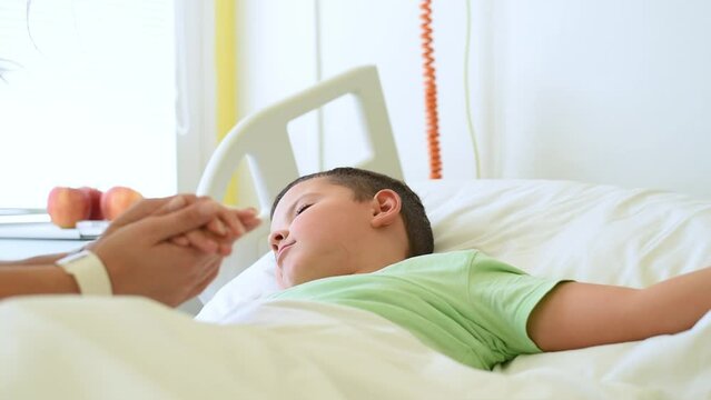 A caring young mother comforts, holds the hand of, and kisses her little son, who is lying in a hospital bed.