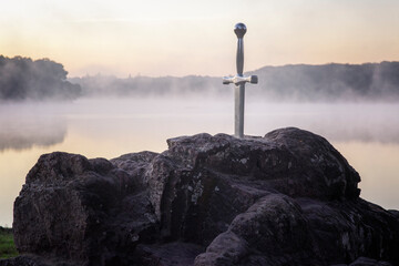 Excalibur l'épée légendaire plantée sur son rocher devant un étang dans la brume du matin au lever du soleil en forêt de Brocéliande - obrazy, fototapety, plakaty