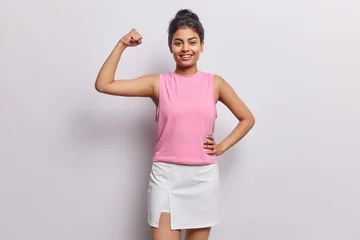 Poster Photo of strong sporty woman raises arm and shows biceps motivates you for doing sport wears pink t shirt and skirt smiles gladfully isolated over white background. You can do it. Healthy lifestyle © Wayhome Studio