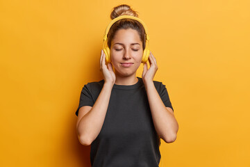 Waist up shot of pleased young woman listens music via headphones keeps eyes closed dressed in...