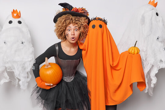 Holiday celebration concept. Indoor photo of young beautiful African american lady in witch costume holding pumpkin standing in centre on white background having party surprised to have many guests