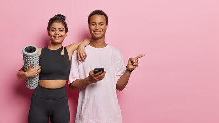Horizontal shot of two sporty woman and man dressed in sportswear carry foam roller for massaging point index finger on copy space for your advertising content isolated over pink background.