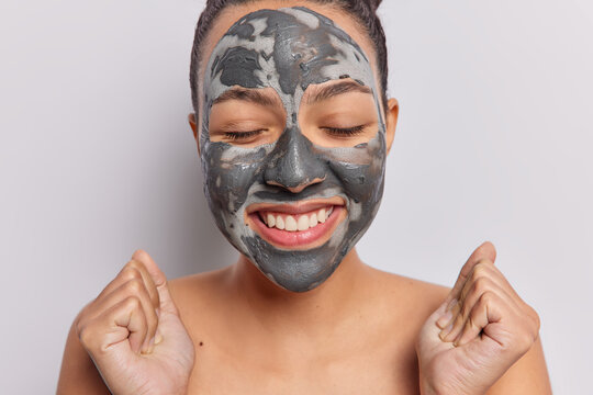 Doing cosmetology procedure concept. Indoor close up of young beautiful excited smiling European lady with closed eyes standing isolated in centre on white background applying clay face mask
