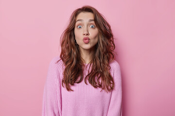 Horizontal shot of lovely dark haired young woman keeps lips folded stares surprised wants to kiss you dressed in knitted jumper isolated over pink background. Human facial expressions concept
