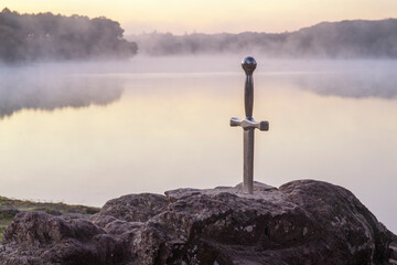 Excalibur l'épée légendaire plantée sur son rocher devant un étang dans la brume du matin au lever du soleil en forêt de Brocéliande - obrazy, fototapety, plakaty