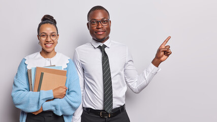 Two ethnic students pose with notepads prepare for examination session dressed in formal clothing indicate at blank space against white background suggests to write your educational promo here