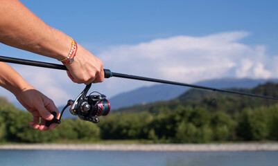 Spinning fishing. Female hands hold a spinning rod against the background of a mountain river, mountains and blue sky in summer on a sunny day, close-up.