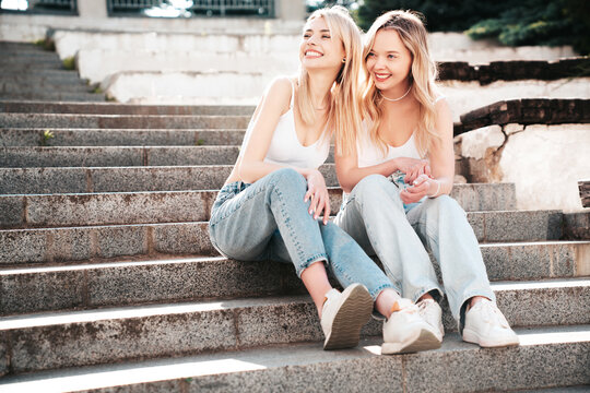 Two young beautiful smiling hipster female in trendy summer white t-shirt and jeans clothes. Carefree women posing in the street. Positive models having fun outdoors. Cheerful and happy. Sit at stairs