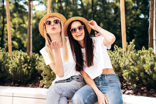 Two young beautiful smiling hipster female in trendy summer white t-shirt and jeans clothes. Carefree women posing in the street. Positive models outdoors. Cheerful and happy. In hat. Give air kiss