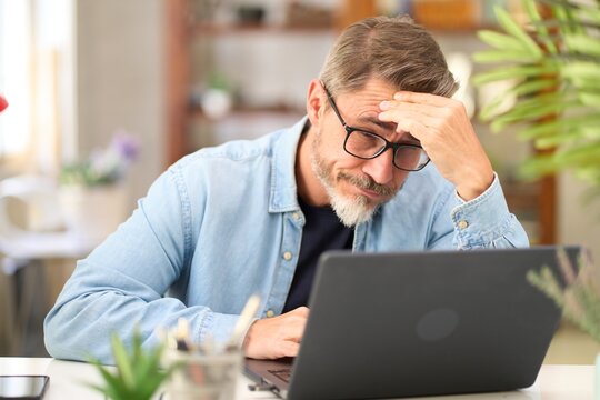 Casual mid adult man with laptop computer at desk in home office, having problem, troubled. Portrait of older gray haired bearded guy thinking. Businessman managing business on internet. &nbsp;