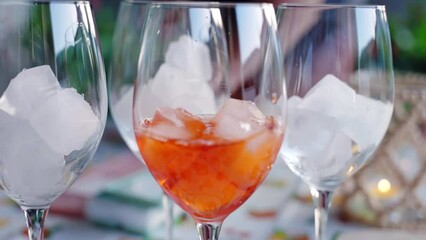 Process of traditional Italian aperol spritz preparation under the open air friends party, bartender pouring aperol into the wine glasses with ice cubes, refreshing aperol spritz cocktail, fizzy