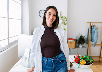 Smiling portrait of young beautiful female nutritionist doctor standing over office workplace....
